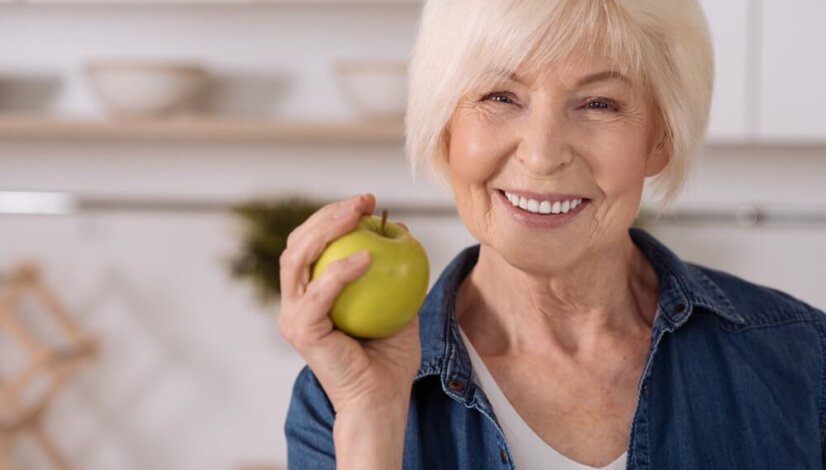 older woman smiling with her dentures as she hold a green apple in her hand