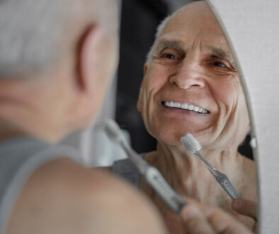 Senior Man Brushing His Dentures In Front Of A Mirror