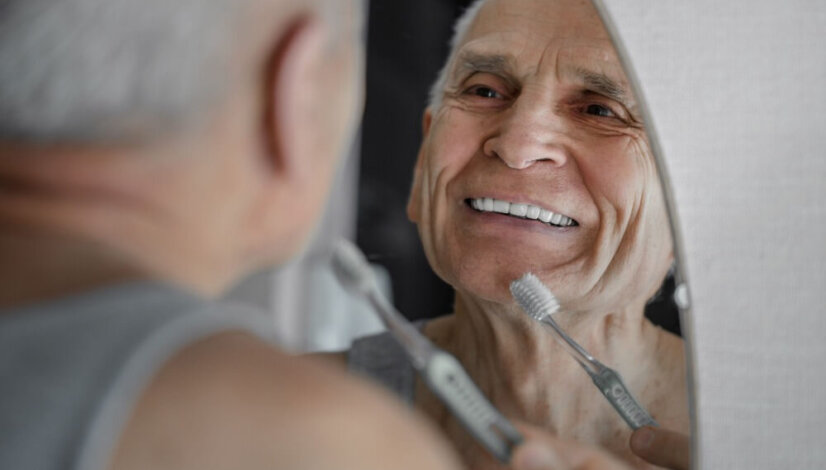 Senior Man Brushing His Dentures In Front Of A Mirror